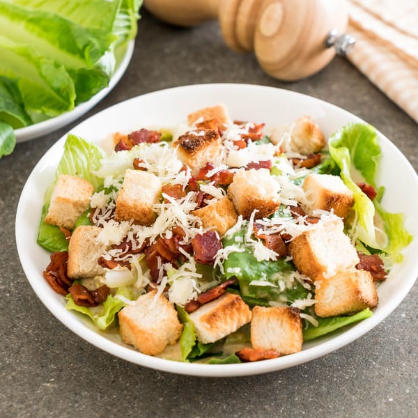 A Caesar salad with romaine lettuce, croutons, bacon bits, and shredded cheese in a white bowl.