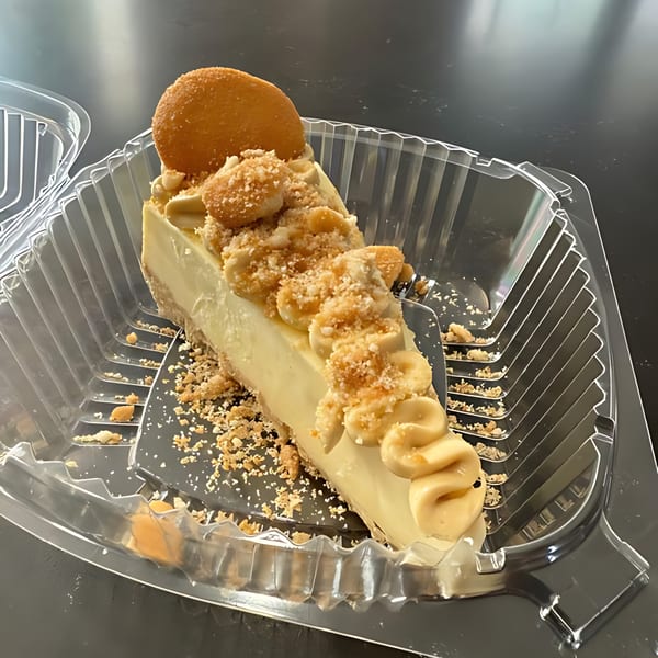 A slice of banana pudding cheesecake with cookie crumbles and a vanilla wafer in a plastic to-go container.