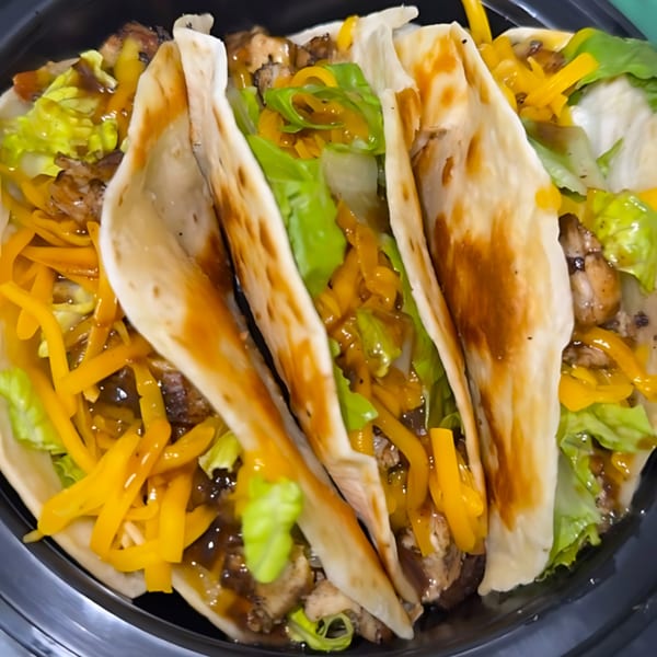 Three soul-food tacos filled with chicken, shredded cheese, lettuce, and sauce in a black container.